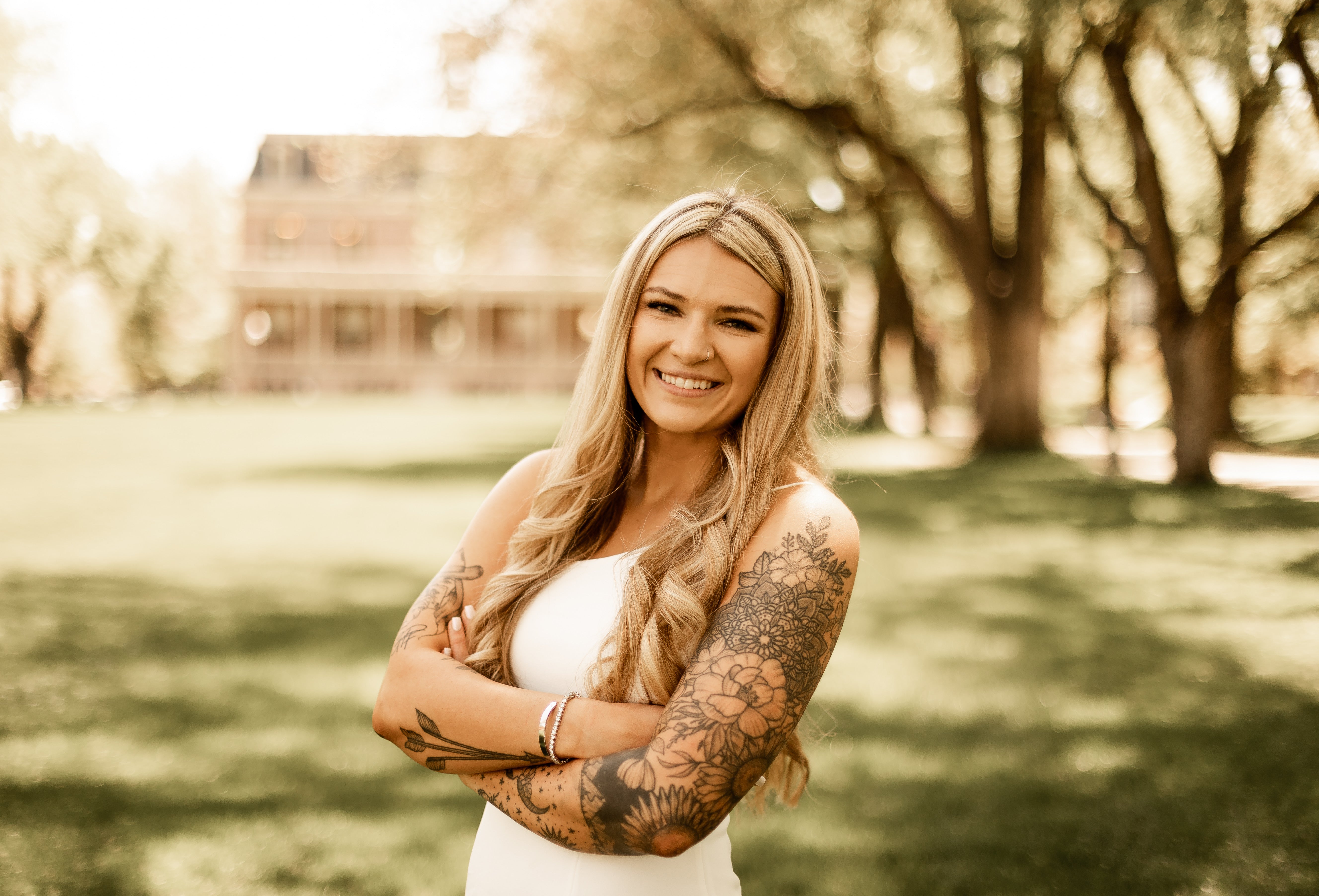 Smiling woman with long blonde hair and visible floral tattoos on both arms, standing with arms crossed in a sunlit grassy park. A large house and rows of trees are softly blurred in the background.