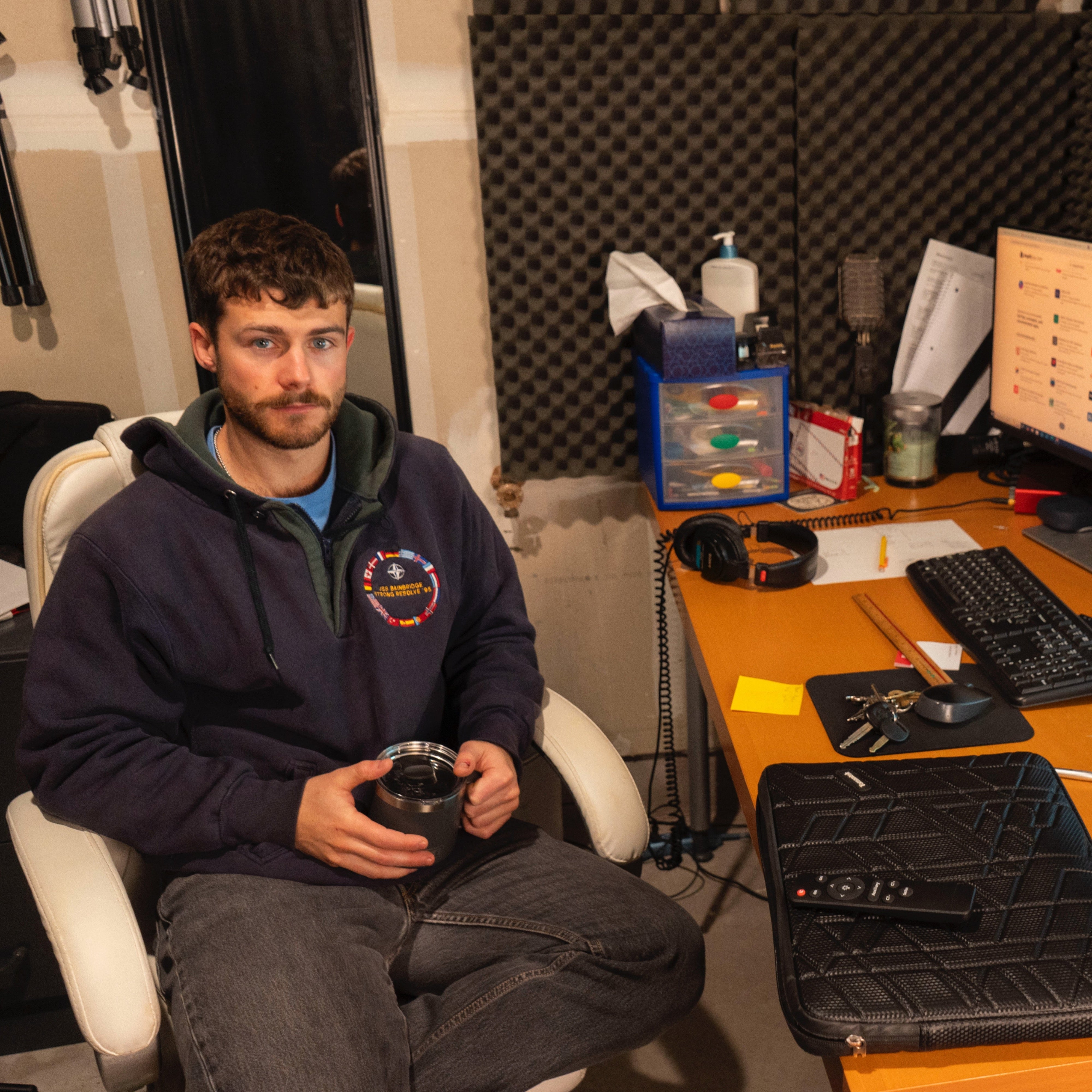 A man sitting in a white office chair holding a drink tumbler in a workspace. He is wearing a navy hoodie with a colorful patch. Behind him are soundproofing foam panels, and in front of him is a desk with a computer monitor, keyboard, headphones, keys, and various office supplies.