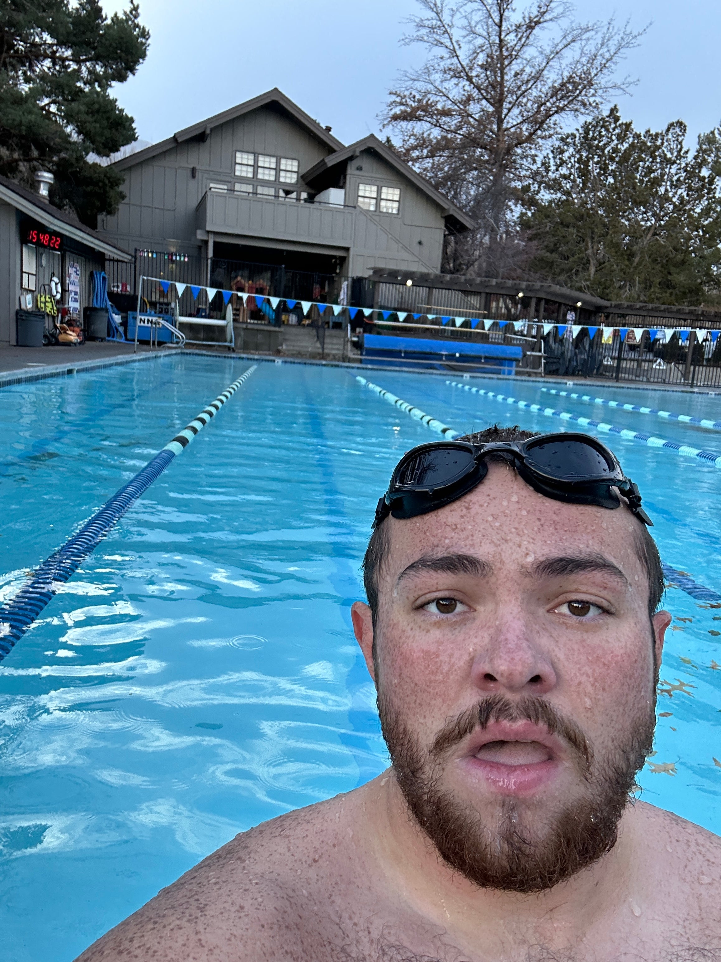 A man with swim goggles resting on his forehead takes a selfie while standing in an outdoor swimming pool. The pool has lane dividers and leads toward a clubhouse building in the background, surrounded by trees. The expression on his face suggests he is catching his breath after a swim.