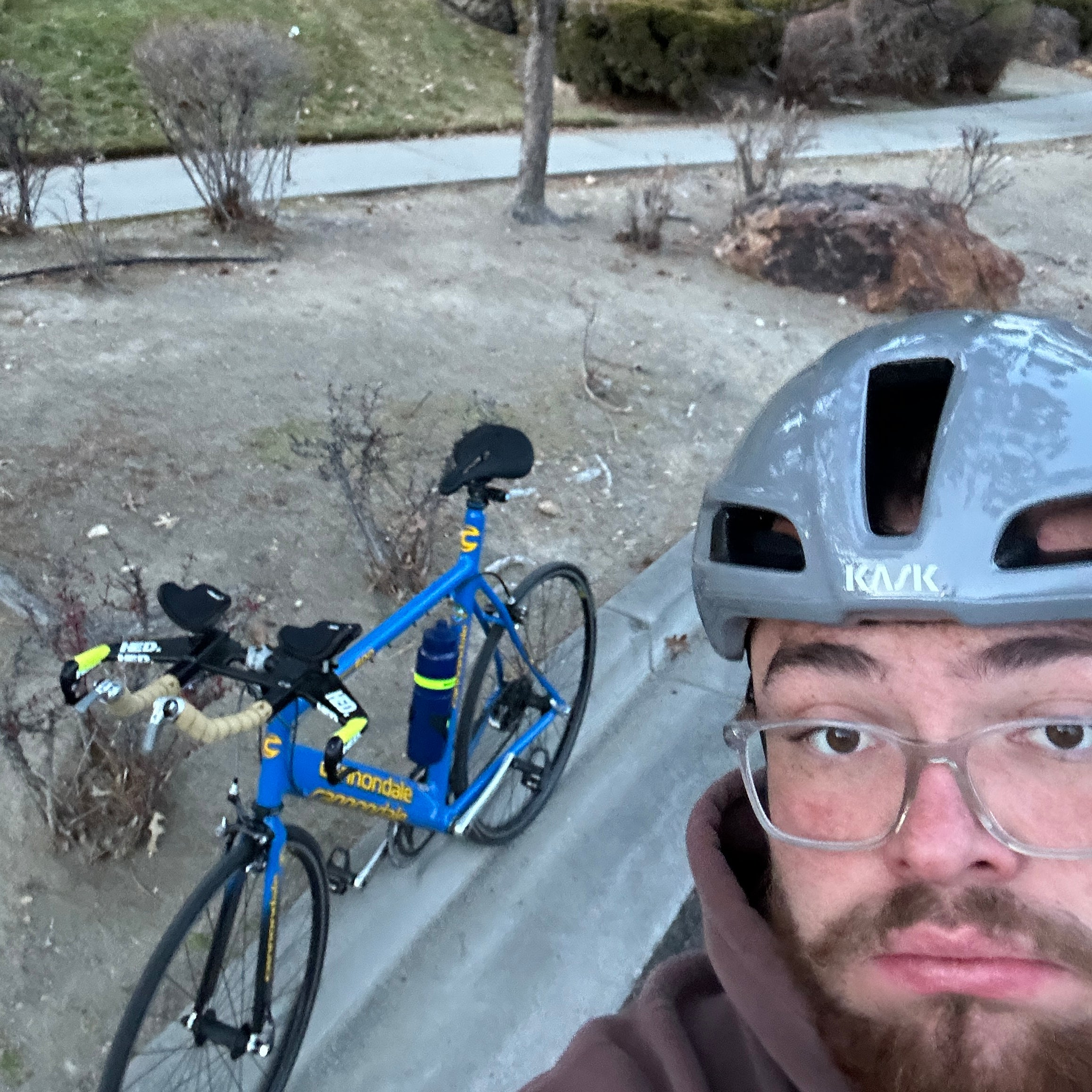 A person wearing a gray KASK bicycle helmet and glasses takes a selfie near the edge of a sidewalk. Behind them is a blue Cannondale road bike with yellow handlebar tape, resting against a curb in a dry, landscaped area with bare shrubs and a dirt patch.