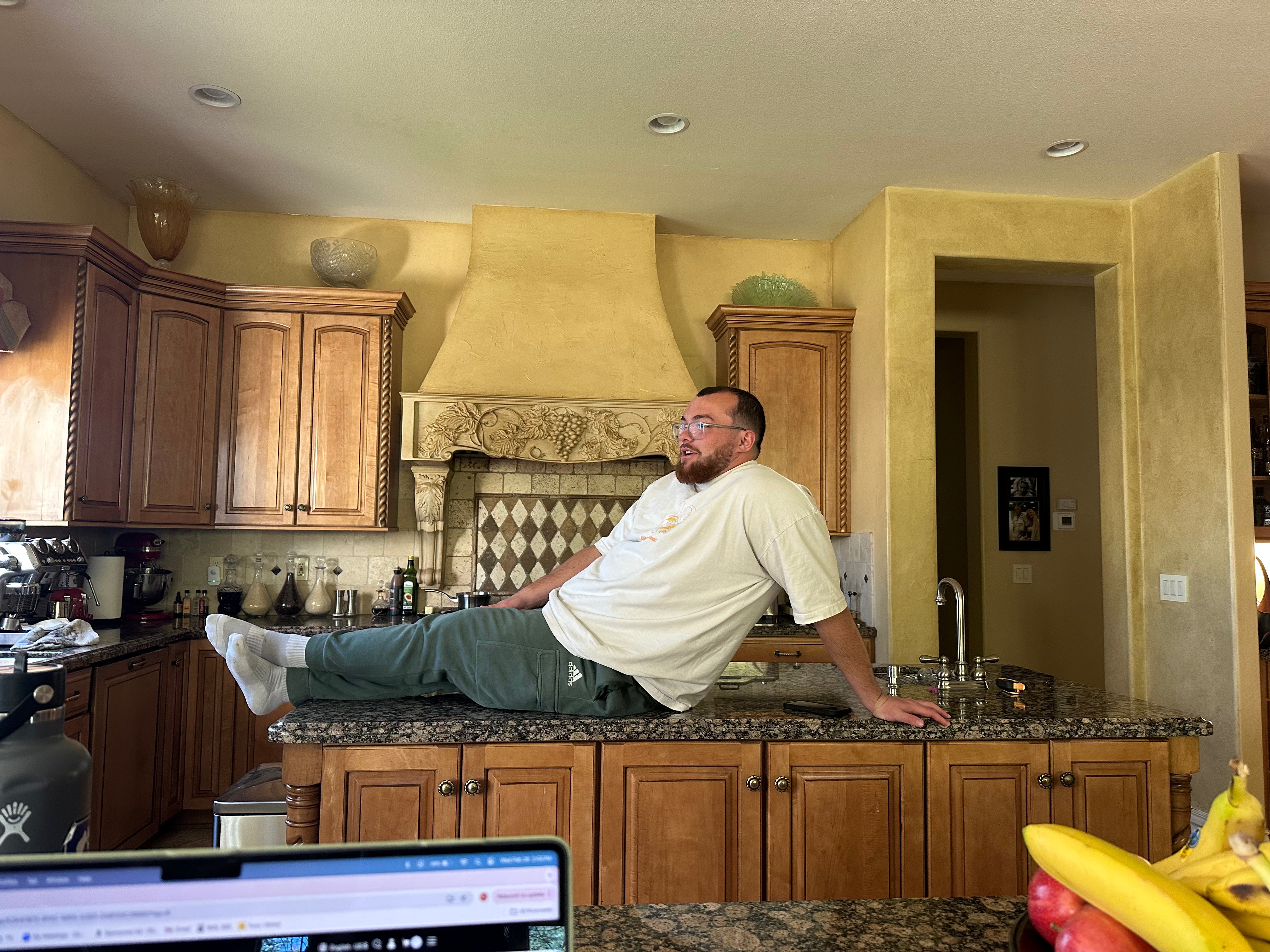 A man lounging on a kitchen counter with one hand supporting him and his legs stretched out. He is wearing a white t-shirt, green sweatpants, and white socks. The kitchen features wooden cabinets, a decorative range hood, and granite countertops. A bowl of bananas and a laptop screen are partially visible in the foreground.