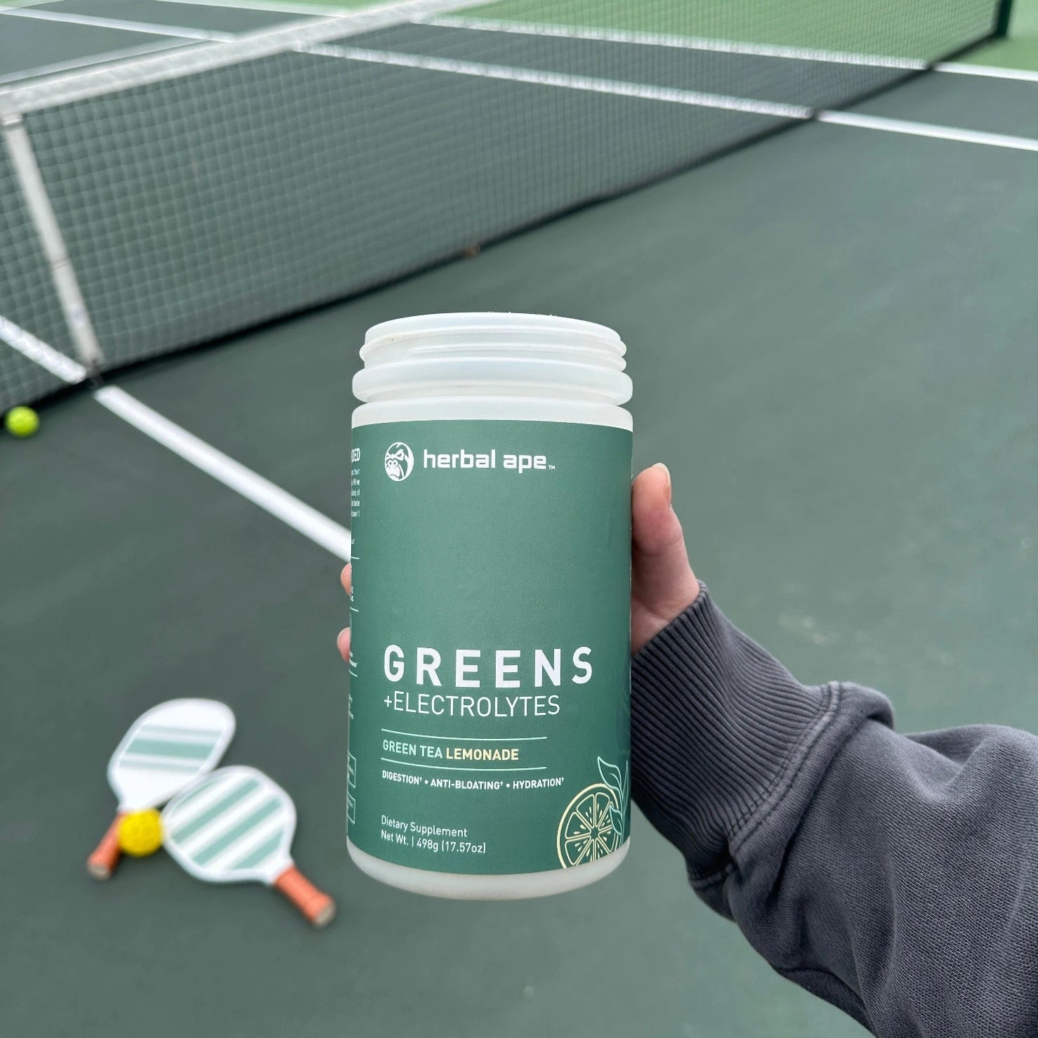 "A person holding a container of Herbal Ape Greens + Electrolytes (Green Tea Lemonade flavor) in front of a pickleball court. Two pickleball paddles and balls are placed on the court surface in the background. The supplement bottle has a matte green label with white and yellow text, emphasizing benefits like digestion, hydration, and anti-bloating.
