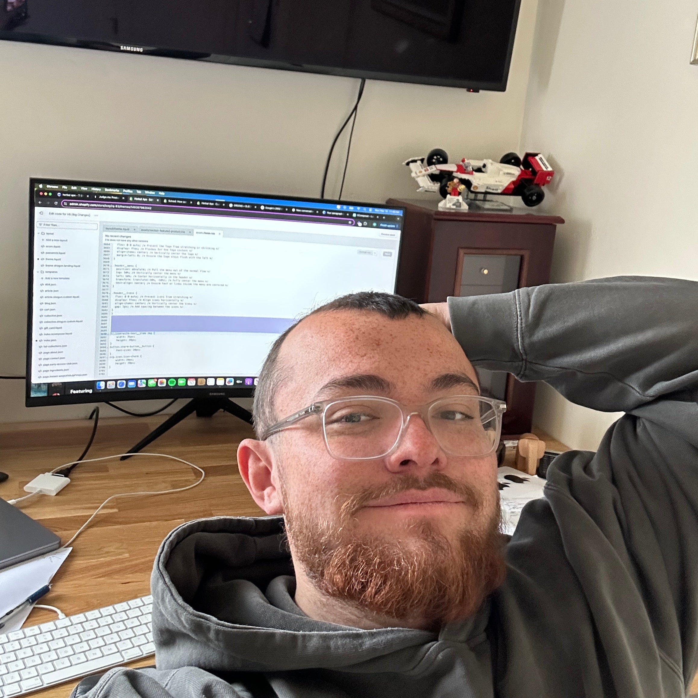 A person wearing glasses and a hoodie taking a relaxed selfie at a desk. Behind them is a computer monitor displaying code or text in a development environment, along with a keyboard, mouse, and LEGO vehicle on a shelf in the background.