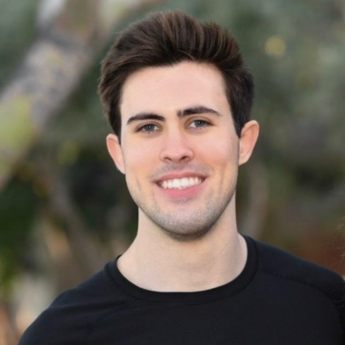 Portrait of a young man smiling outdoors, wearing a black shirt. He has short, styled dark hair and is standing in front of a blurred background of trees and greenery.