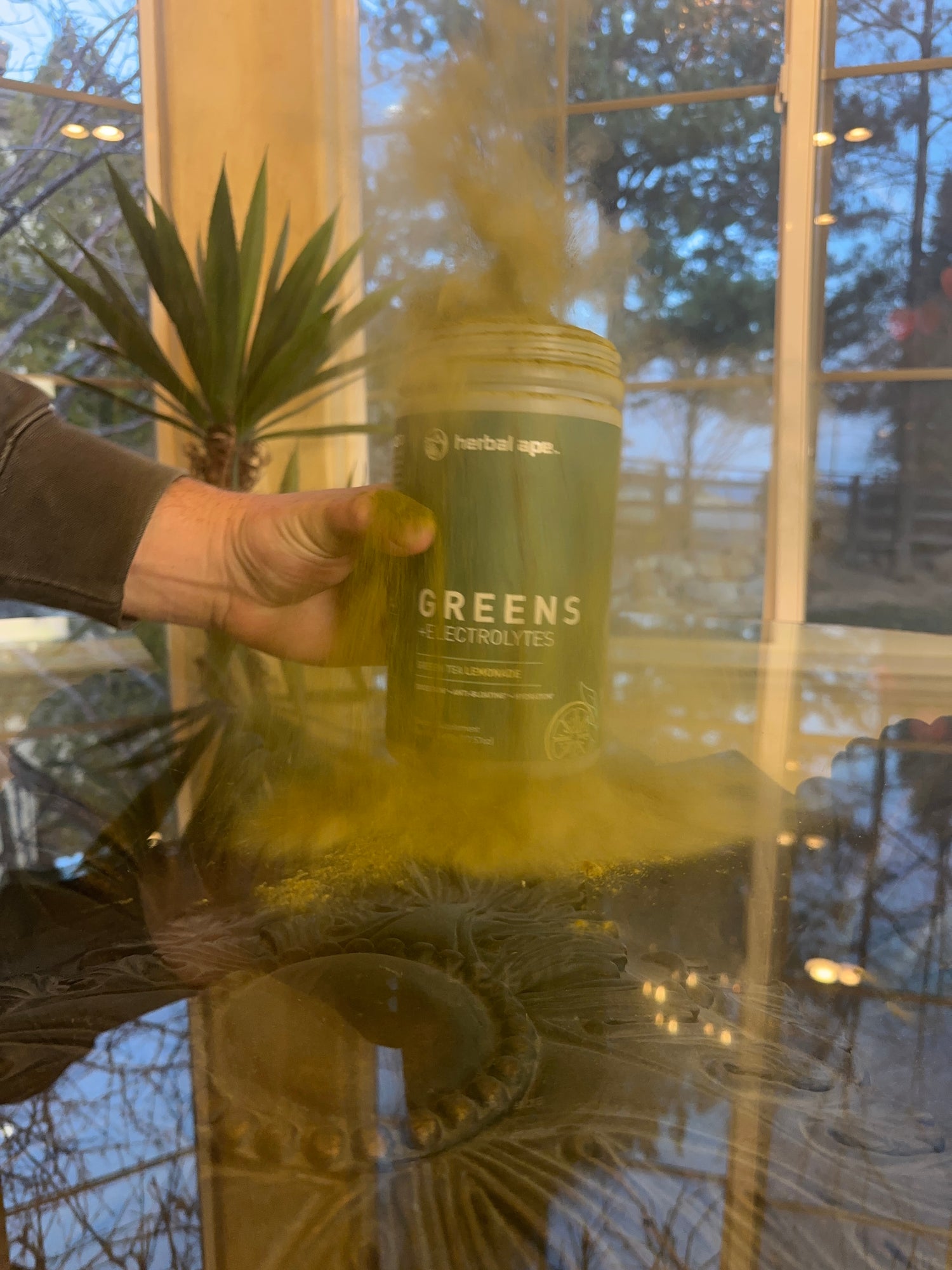 A hand holding a container of Herbal Ape Greens + Electrolytes powder, with green powder dramatically bursting into the air. The scene is set indoors near a window with plants and trees visible in the background.