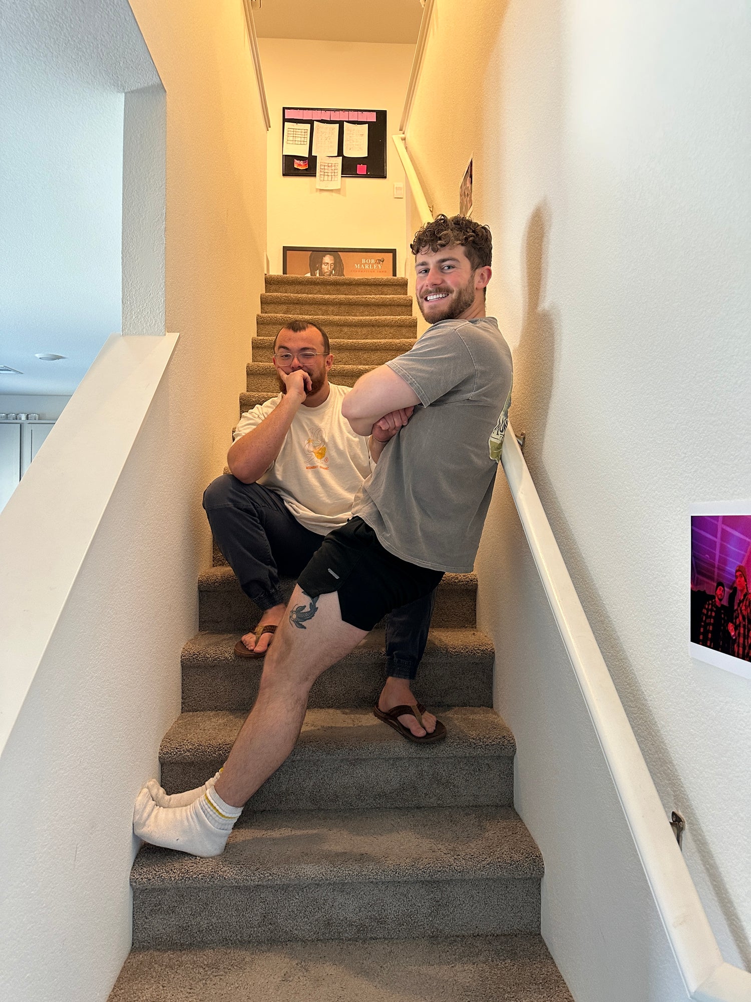 Two men posing playfully on a carpeted staircase inside a home. One is crouching on a step with his hand on his chin, wearing a white shirt and flip-flops. The other is smiling with arms crossed, leaning back on a lower step, wearing a gray shirt, black shorts, and white socks.