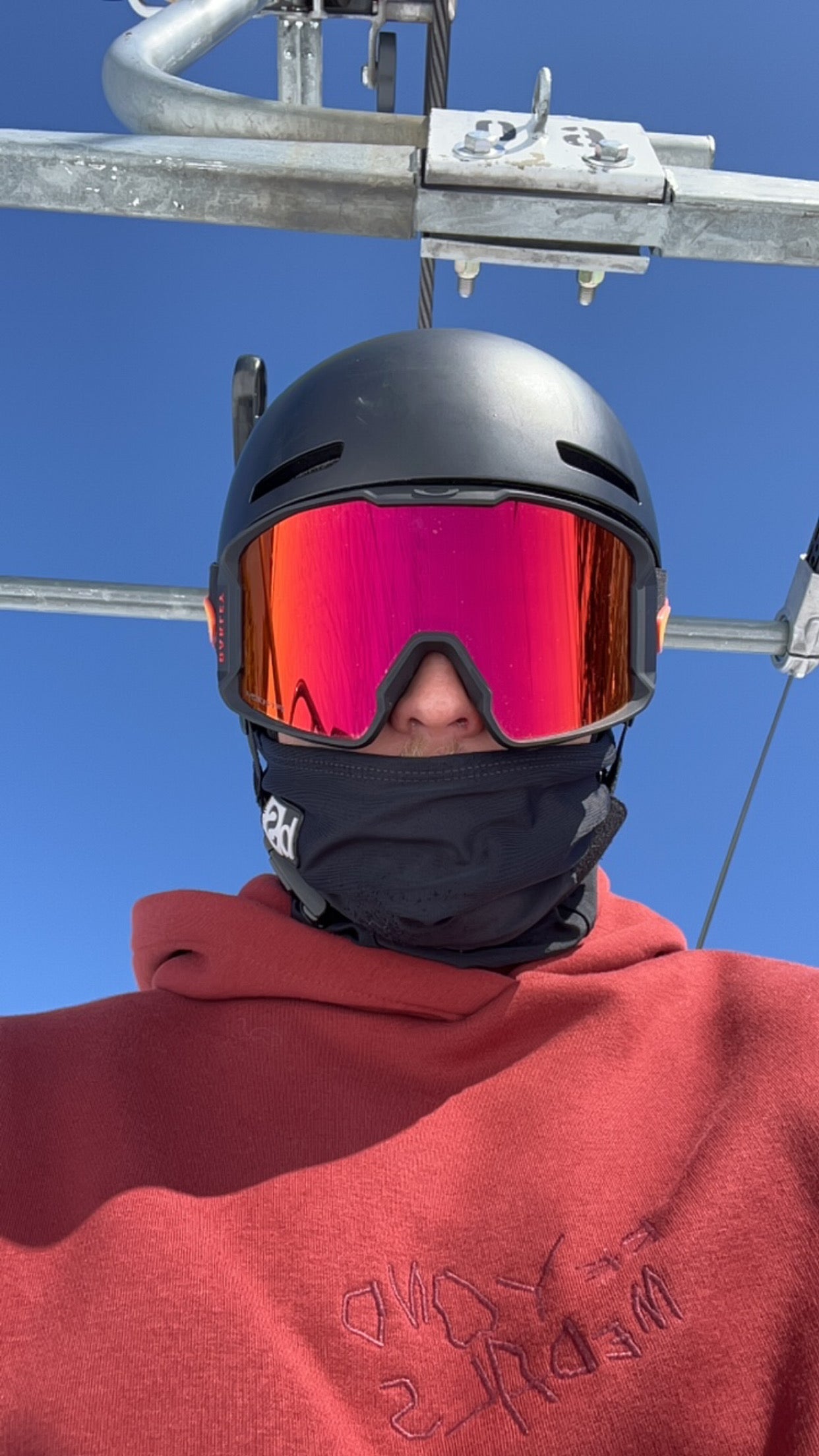 Person wearing a black ski helmet, reflective red ski goggles, and a face covering, sitting on a ski lift under a clear blue sky. They are dressed in a red hoodie with darker red text partially visible on the chest.