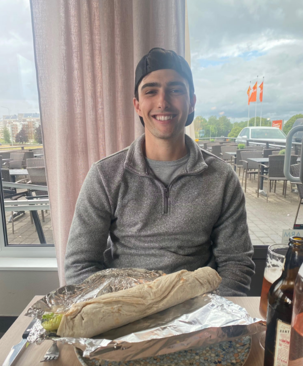 A smiling young man sitting at an indoor restaurant table with a large wrapped sandwich in front of him. He is wearing a dark baseball cap backward and a gray quarter-zip sweater. Beer bottles and utensils are also visible on the table, with patio seating and orange flags seen in the background.