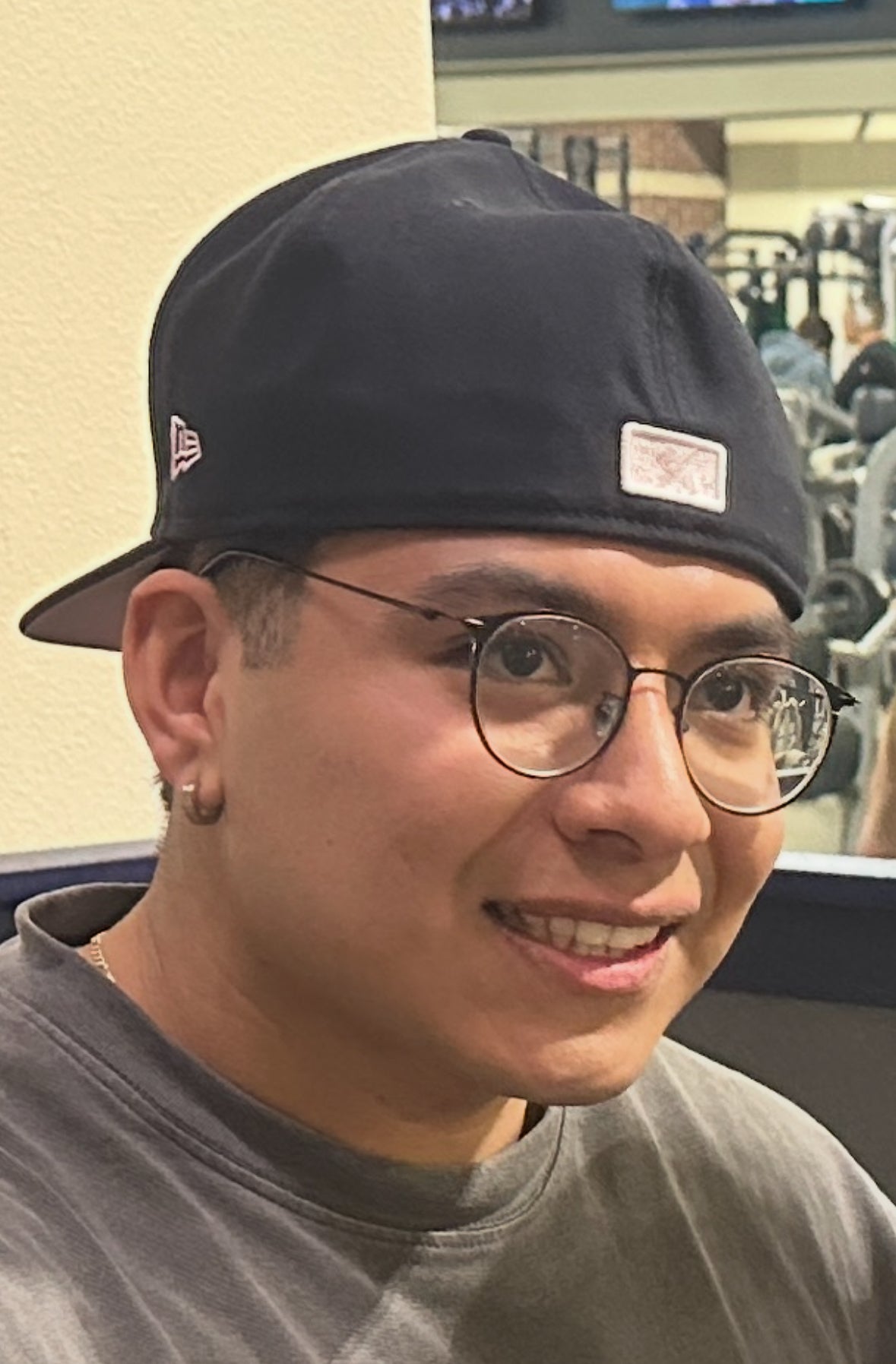 Close-up of a smiling young man wearing glasses, a dark t-shirt, and a backward black baseball cap. He has a small hoop earring in one ear and is indoors, possibly in a gym setting.