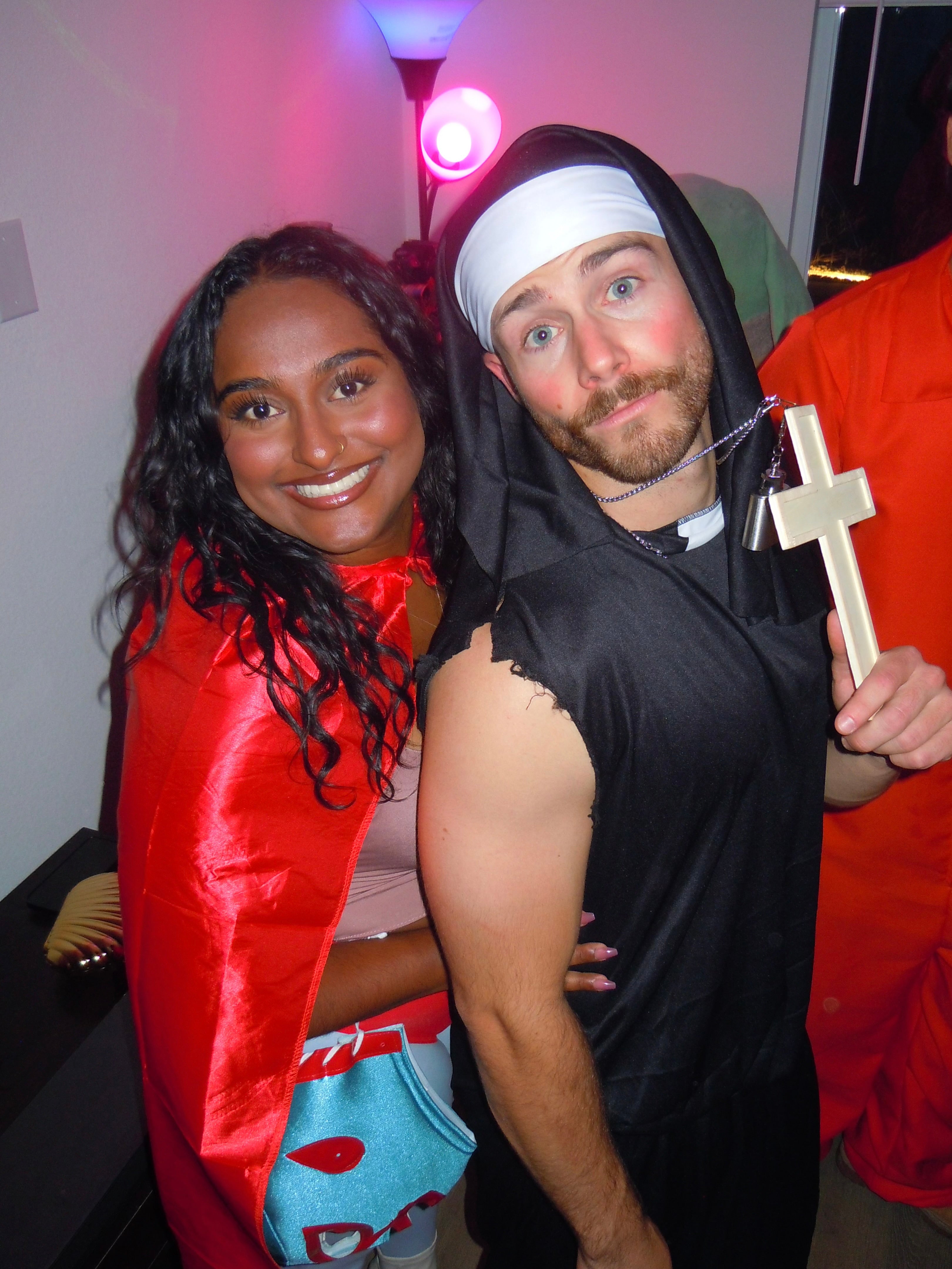 Two people posing together in costumes at a party. The woman on the left is dressed in a red cape and holding a blue cartoon-themed bag, smiling at the camera. The man on the right is dressed in a black nun costume with a white headband and holding a wooden cross, with a playful expression.