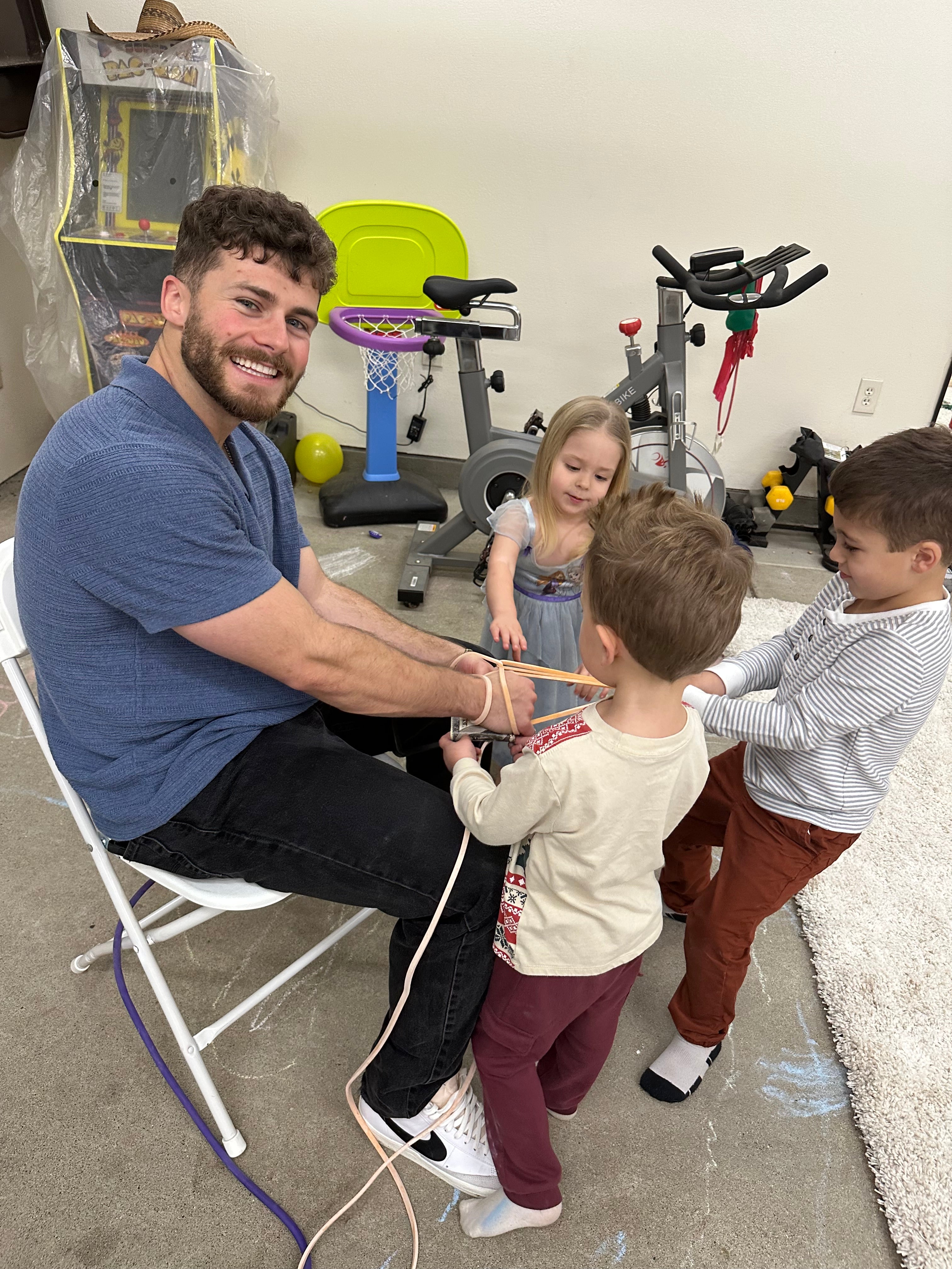 A smiling man seated on a white folding chair with three young children playfully tying ropes around his arms. The scene takes place in a home gym or playroom with exercise equipment, a small basketball hoop, and toys visible in the background.