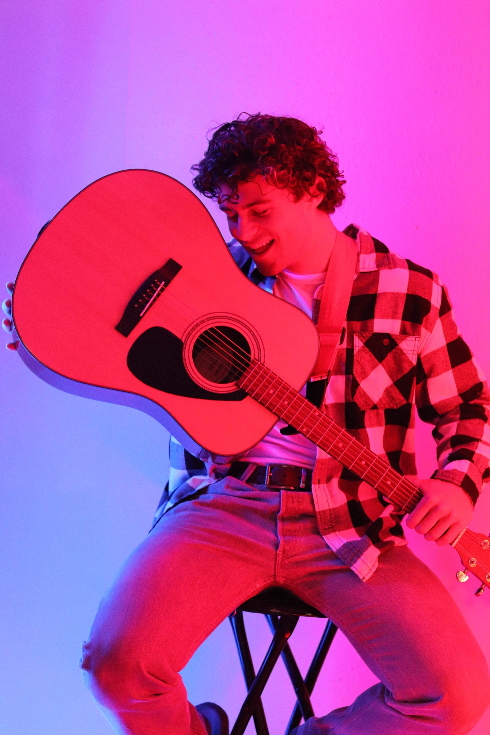 A young man sitting on a stool and smiling while holding an acoustic guitar. He is wearing a black and white checkered flannel shirt and jeans. The scene is lit with vibrant pink and blue lighting, creating a colorful, artistic atmosphere.