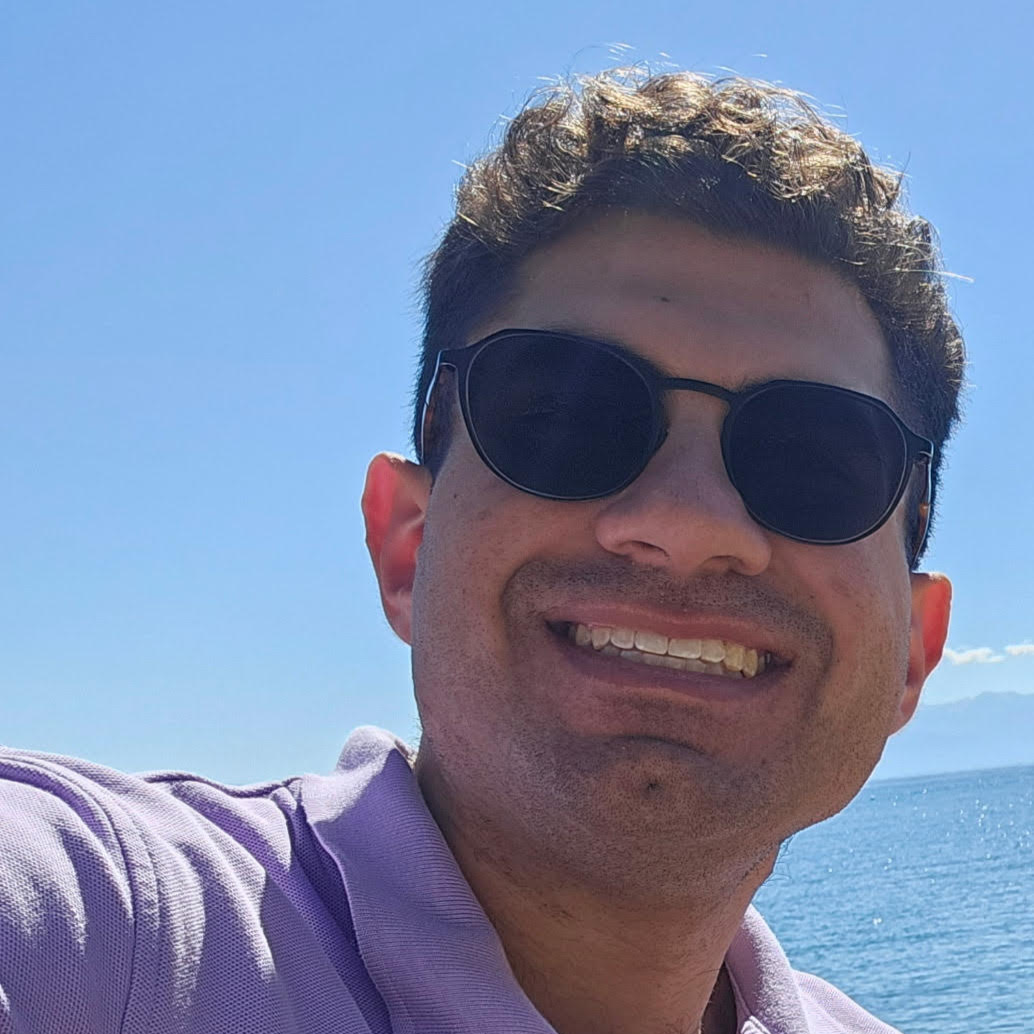 Close-up selfie of a man wearing dark sunglasses and a light purple shirt, smiling in front of a sunny ocean or lake backdrop with blue skies and distant mountains.