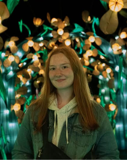 Young woman with long red hair smiling slightly, standing in front of a decorative light display featuring glowing flowers and green stems. She is wearing a denim jacket over a light hoodie and has a crossbody bag.