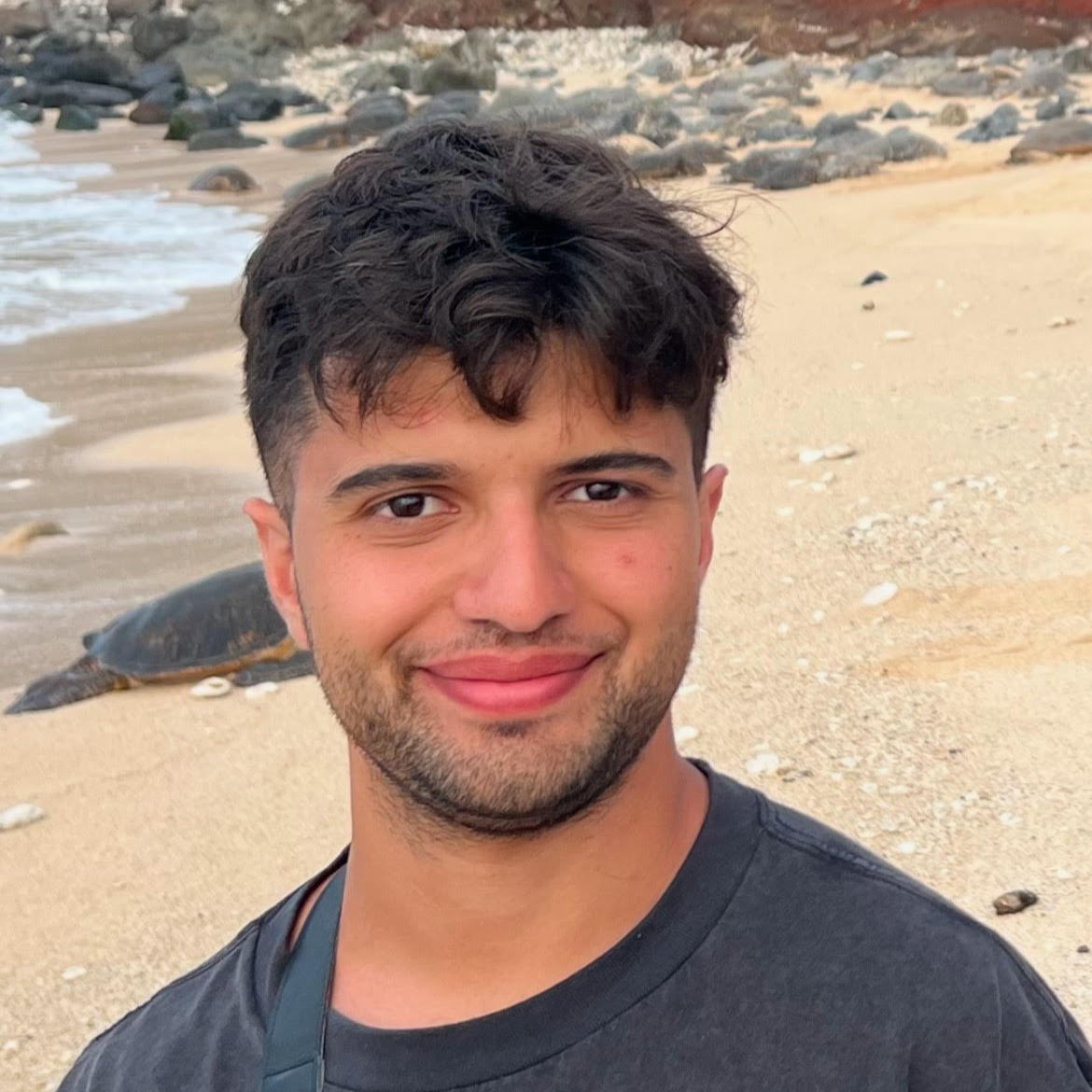 Smiling man standing on a sandy beach with ocean waves and rocks in the background. A sea turtle is visible resting on the shore behind him. He is wearing a dark t-shirt and has short, tousled hair and a trimmed beard.